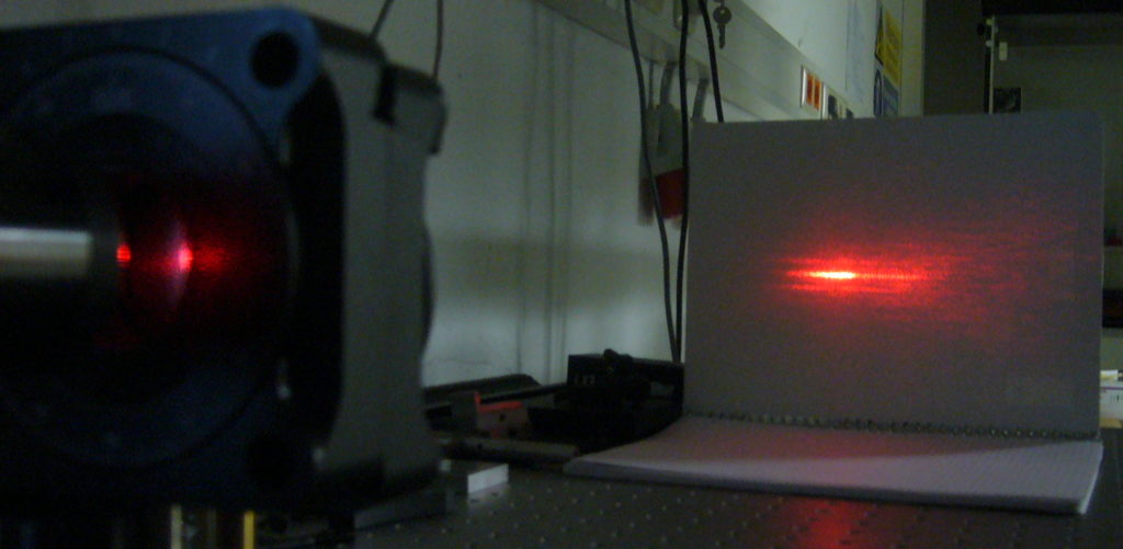 Ein Laser wird (von links kommend) durch ein feinmaschiges Gitter geschickt um am Ort des Blatt Papiers ein Interferenzmuster zu erzeugen.